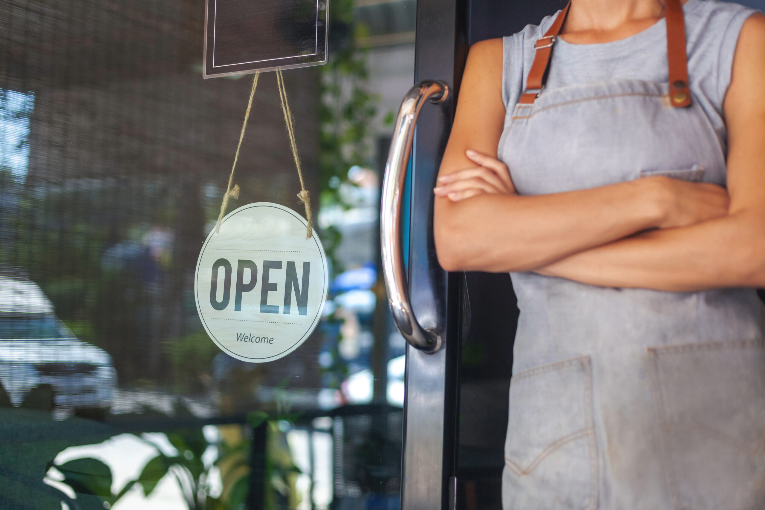 A small business owner in an apron stands beside a glass door with an “Open” sign, representing local shops in Franklin, TN.